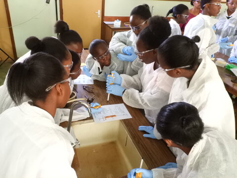 An instructor holding a yellow micropipette showing a group of high school students in lab coats how to use it